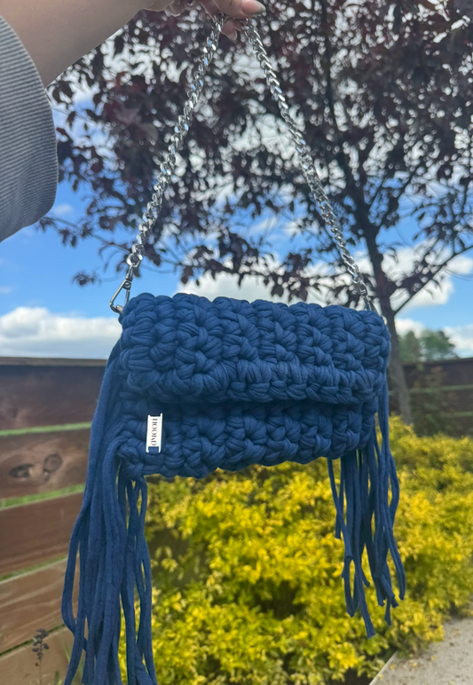 Blue crochet bag with tassels held against a natural background with trees and sky.